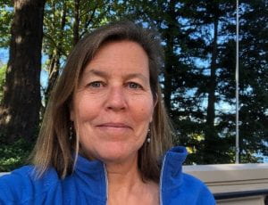 Ginny Boradhurst. Ginny is smiling, has shoulder-length brown hair, and is wearing a blue fleece zip-up. Trees with broken sunshine and blue sky behind her.