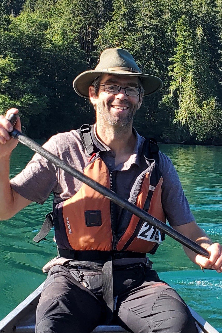 T. Abe Lloyd is in a canoe. He is smiling and holding a paddle. He is wearing a hat, glasses, long pants and has on a life vest.
