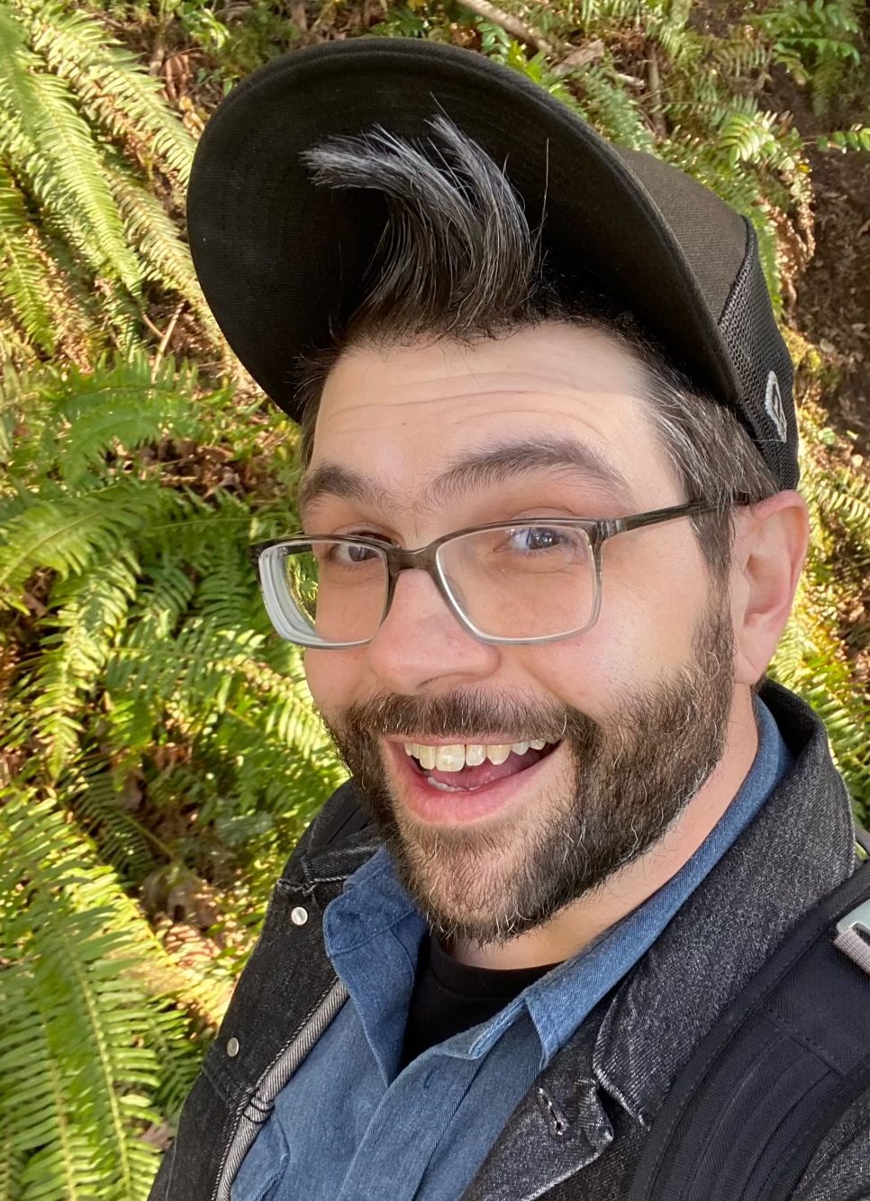Josh is smiling, has a full beard, is wearing a hat and glasses. There are ferns and tress in the background.