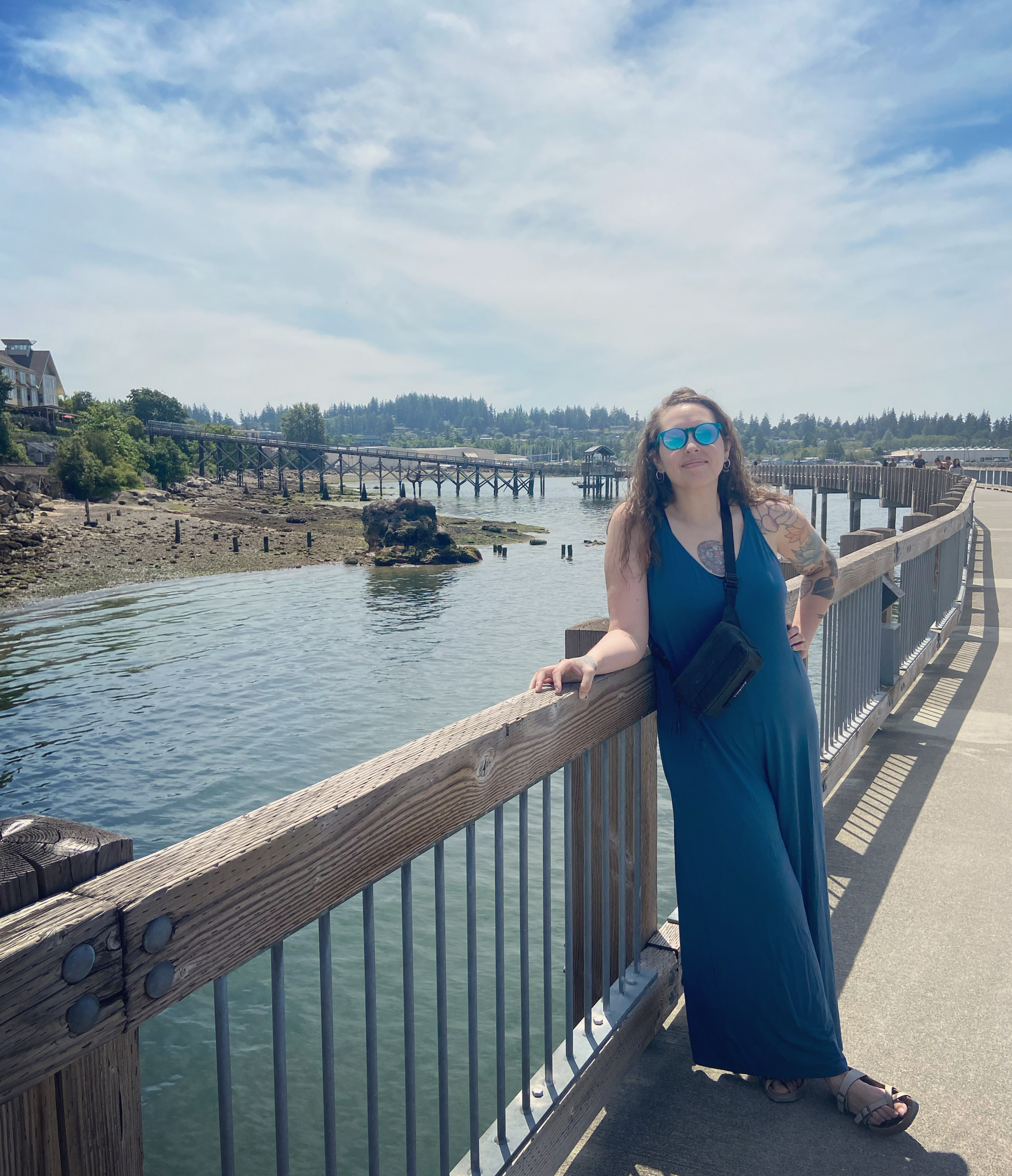 Theresa is leaning against the railing of a walkway over the water. She is wearing a blue jumper and sunglasses. The sky is light blue with wispy clouds.