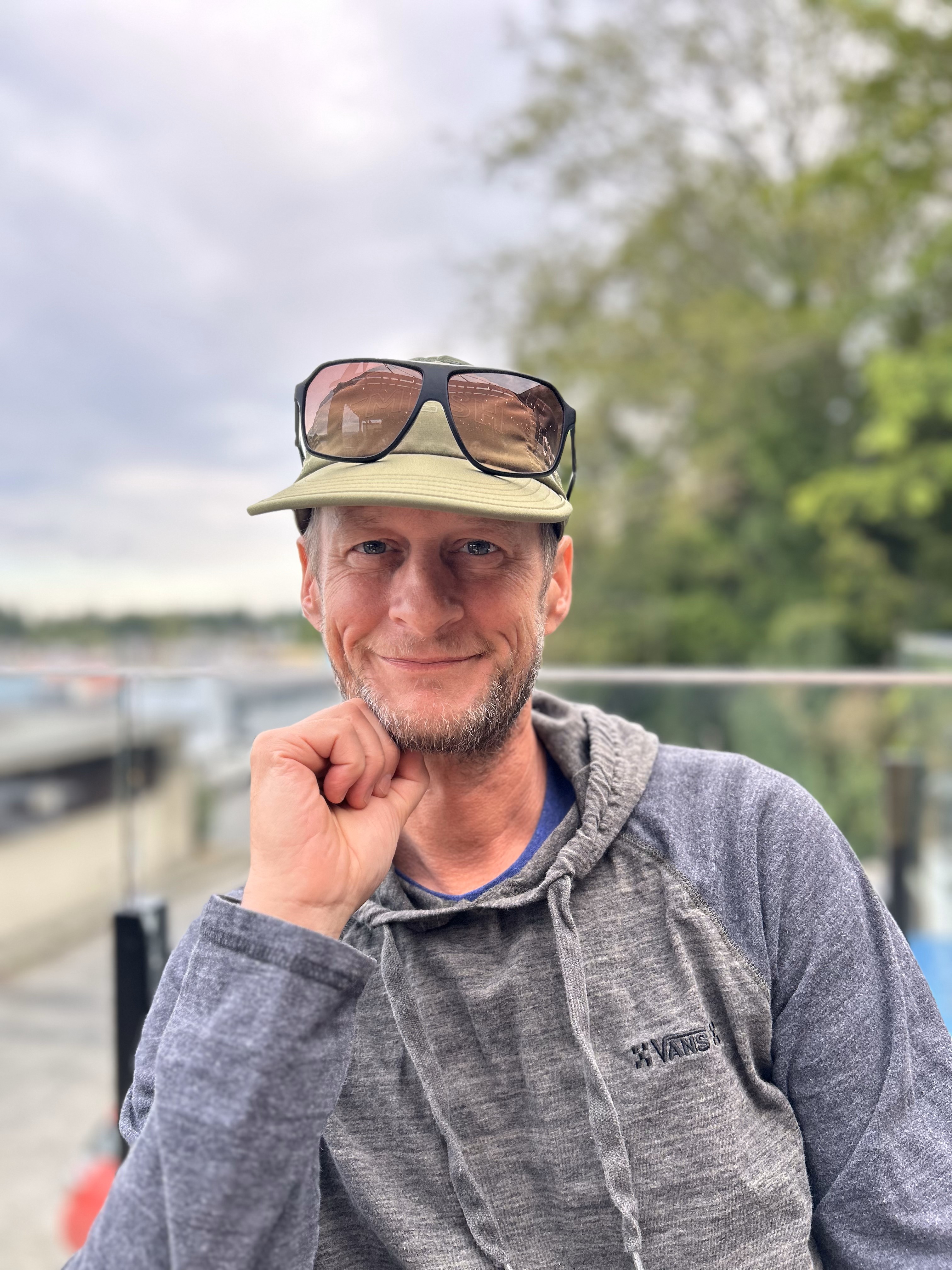 David is sitting in a chair on a dock at the water's edge. He is smiling at the camera with his right hand under his chin. He is wearing a hoodie and a hat with sunglasses on top of his hat.