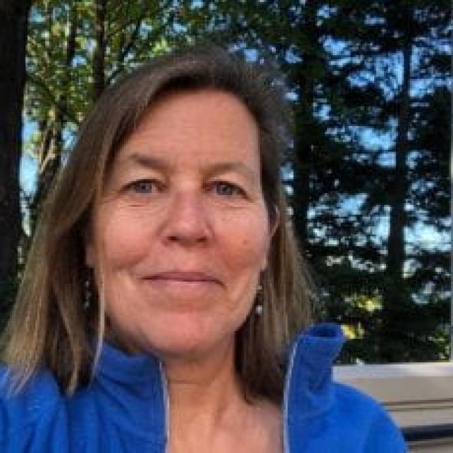 Ginny Boradhurst. Ginny is smiling, has shoulder-length brown hair, and is wearing a blue fleece zip-up. Trees with broken sunshine and blue sky behind her.