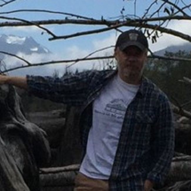James is standing with Mount St. Helens in the background. He is wearing a t-shirt with a long sleeve flannel, a ball cap, and glasses.