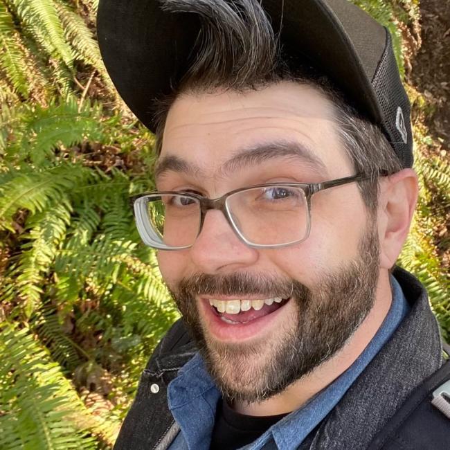 Josh is smiling, has a full beard, is wearing a hat and glasses. There are ferns and tress in the background.