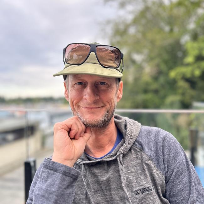 David is sitting in a chair on a dock at the water's edge. He is smiling at the camera with his right hand under his chin. He is wearing a hoodie and a hat with sunglasses on top of his hat.